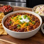 A bowl of chili topped with sour cream, cheddar cheese, and green onions, surrounded by tortilla chips, salsa, coleslaw, and brownies on a wooden board.