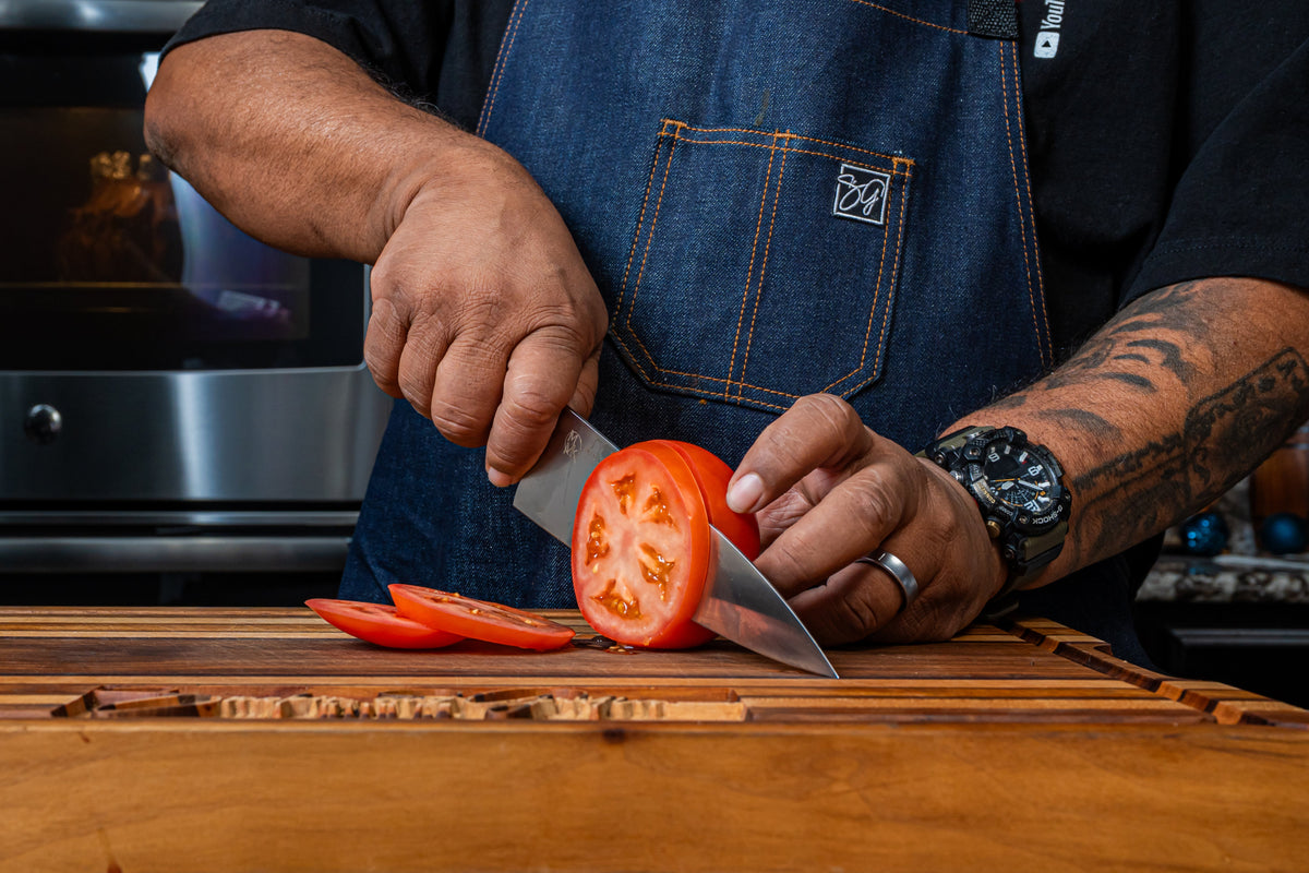 AB slicing a tomato with his red-handled signature chef knife on a wooden cutting board.