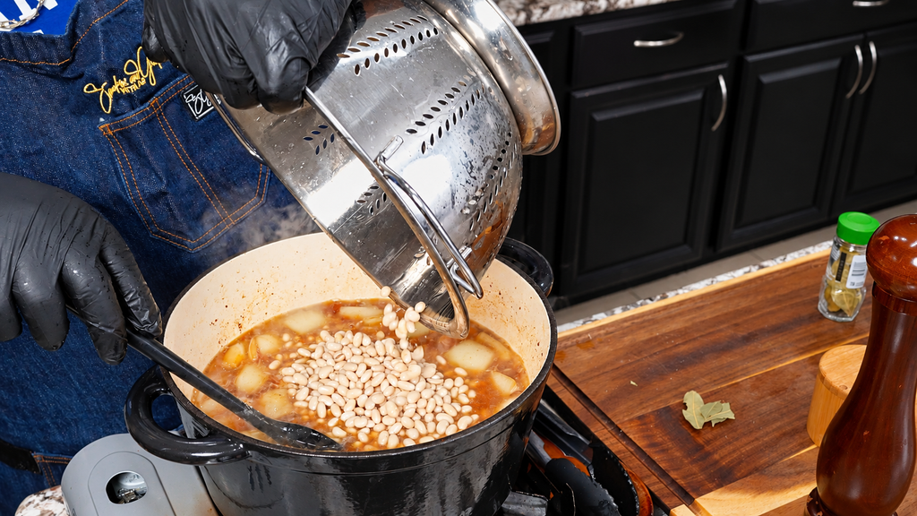 A person wearing black gloves pours white beans from a metal colander into a pot of stew on a stove.