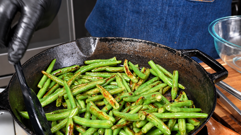 A person wearing black gloves uses a spatula to stir dirty green beans and potatoes in a cast-iron skillet.