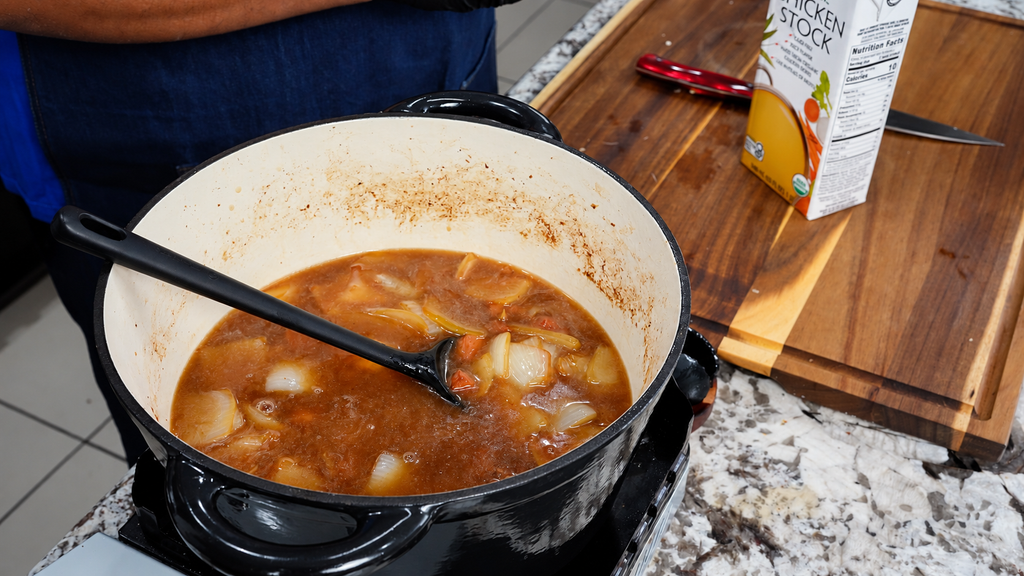 A black enameled cast iron pot filled with simmering brown broth, onion chunks, and carrots, with a serving spoon resting inside and a carton of chicken stock on the wooden cutting board nearby.