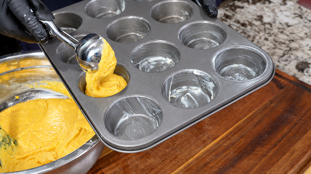 Cornbread batter being scooped into the cups of a greased muffin tin.