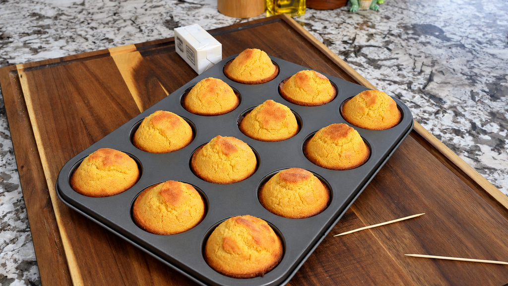 Golden baked cornbread muffins resting in a muffin tin on a wooden board.