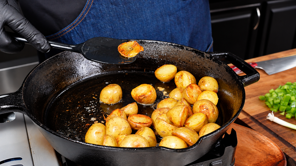 A gloved hand uses a spatula to lift a golden-brown potato piece from a cast iron skillet filled with dirty green beans and potatoes.
