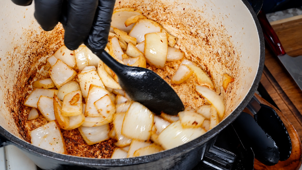 A person wearing black gloves uses a black spatula to stir golden-brown sautéed onion chunks in a Dutch oven.