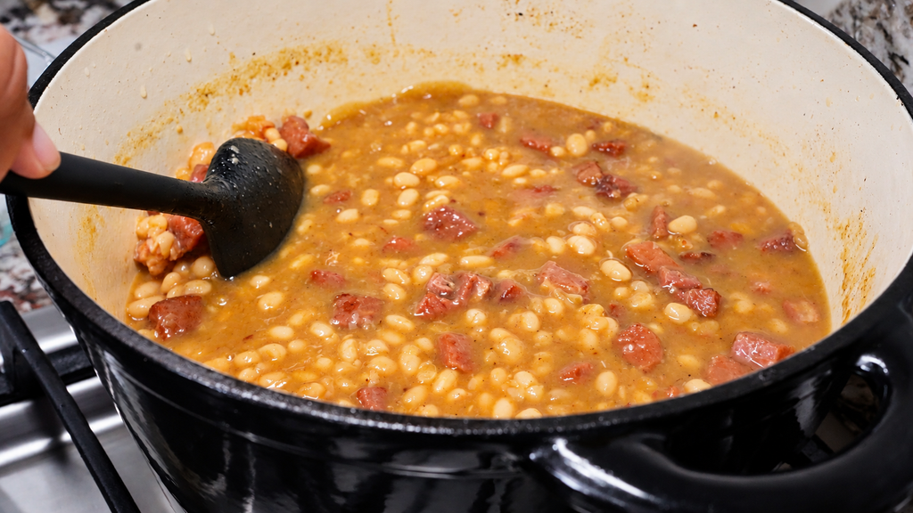 A hand uses a black spatula to stir baked beans with chunks of meat in a large enameled cast iron pot on a stove.